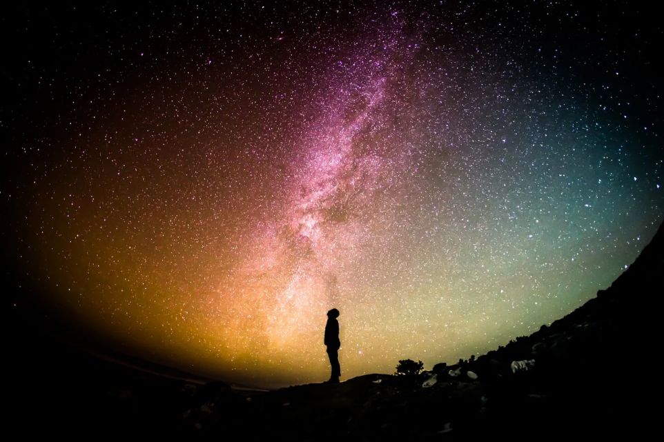 Silhouette eines Mannes mit Blick nach oben auf eine vollen Sternenhimmel. Dieser weist eine starke Krümmung auf, zeigt im Inneren die Milchstraße und eine Farbverlauf von rot bis grün.