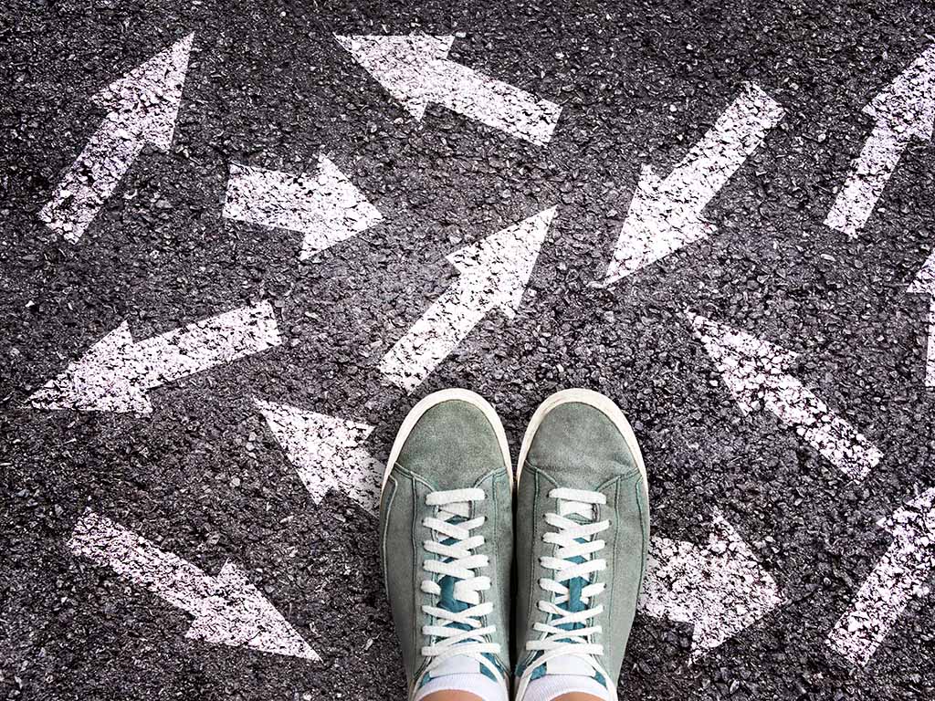 Auf dem Asphaltboden sind Pfeile in verschiedene Richtungen zu sehen und ein Paar Turnschuhe. Fragestellung: Welchen Weg gehen?