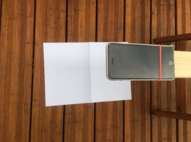 Nahaufnahme von einem Smartphone an einer Holzlatte welches auf ein Blatt Papier am Boden gerichtet ist