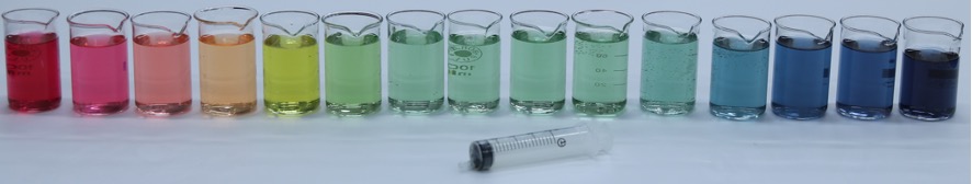 Das Bild zeigt 15 in einer Reihe aufgestellte Bechergläser, deren Flüssigkeit verschiedene Farben aufweist. Der Farbverlauf beginnt auf der linken Seite mit einem kräftigen Rot, gefolgt von Rosa, Orange und Gelb. Weiter in der Mitte gehen die Farben in ein Hellgrün und Grün über. Danach wird die Flüssigkeit bläulicher, beginnend mit einem Hellblau, das sich schließlich in ein kräftigeres Blau und Dunkelblau verwandelt. Vor den Bechergläsern liegt eine leere Spritze.