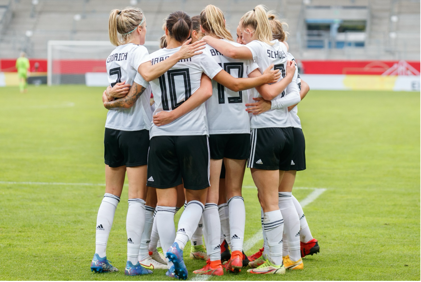 Auf dem Foto stehen mehrere Fußballspielerinnen in weißen Trikots und schwarzen Hosen der deutschen Nationalmannschaft in einem engen Kreis und umarmen sich. Sie befinden sich auf einem Rasenplatz in einem Stadion, dessen Tribüne im Hintergrund zu sehen ist.