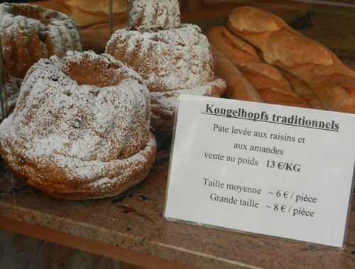 Ein Schaufenster mit traditionellen Kougelhopfs, bestäubt mit Puderzucker. Daneben steht ein Schild mit Informationen: Kougelhops traditionnels - Pâte levée aux raisins et aux amandes, vente au poids 13 €/KG. Taille moyenne - 6 €/pièce, Grande taille - 8 €/pièce. Im Hintergrund sind Baguettes zu sehen.