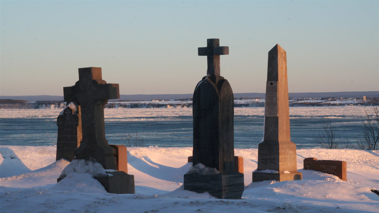 Schneebedeckte Grabsteine und Kreuze vor einem gefrorenen Fluss, mit einer winterlichen Landschaft im Hintergrund