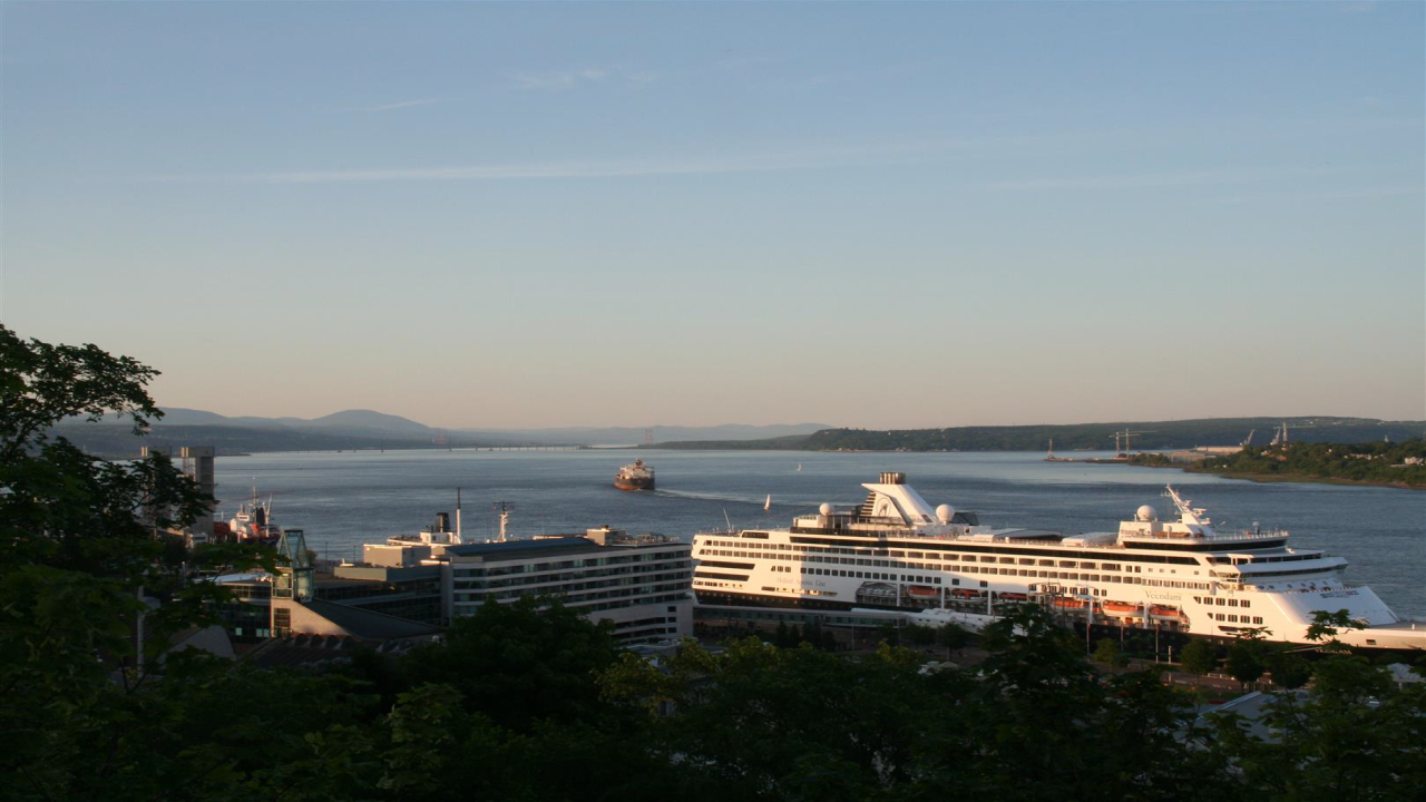 Ein großes Kreuzfahrtschiff liegt in einem Hafen, umgeben von ruhigem Wasser und grünen Hügeln im Hintergrund