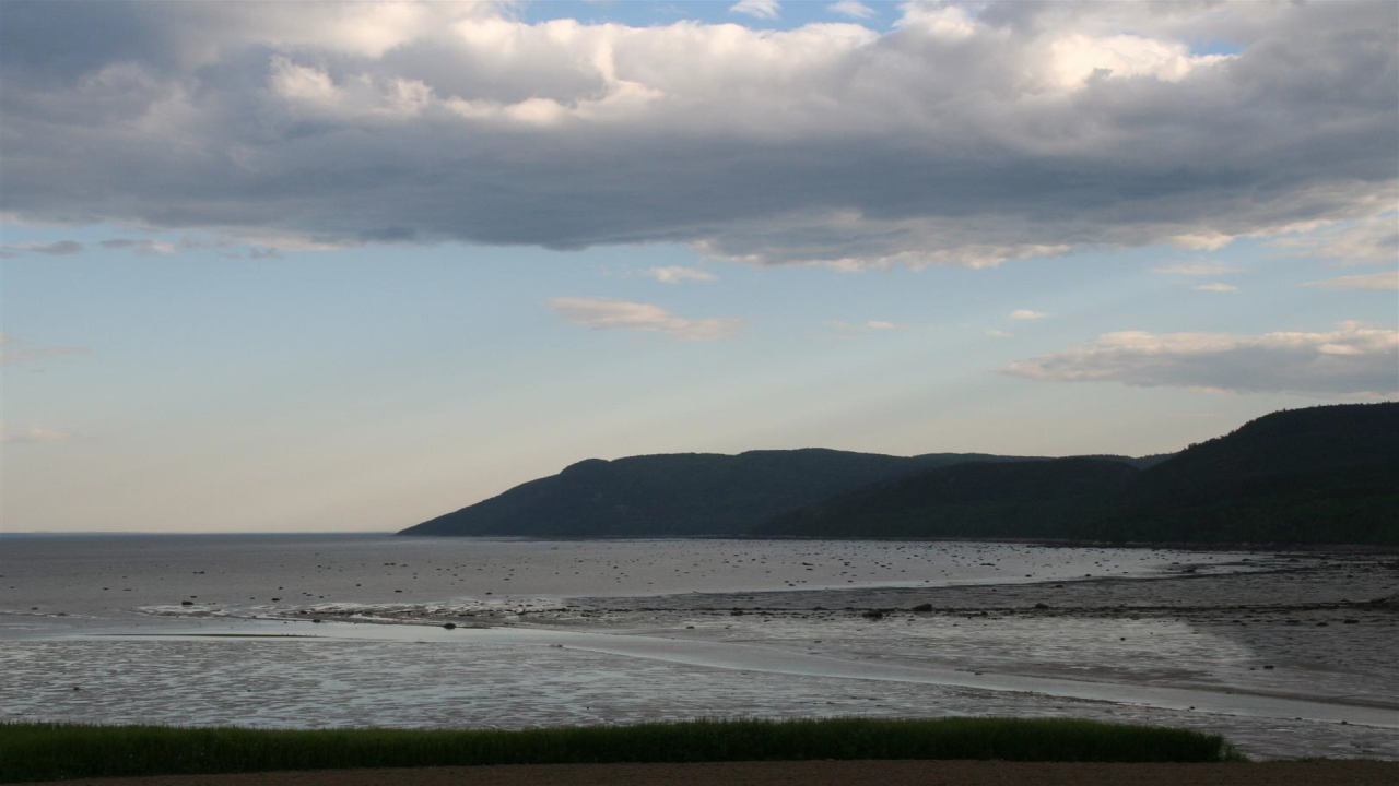 Küstenlandschaft mit einem ruhigen Meer, sandigen Ufern und bewaldeten Hügeln im Hintergrund unter einem bewölkten Himmel