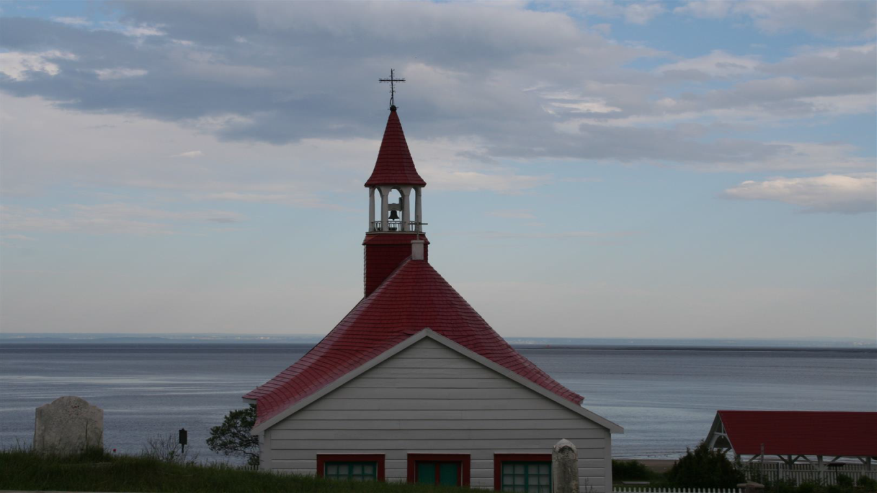 Eine kleine Kirche mit rotem Dach und Glockenturm steht vor einem ruhigen Gewässer, unter einem teils bewölkten Himmel