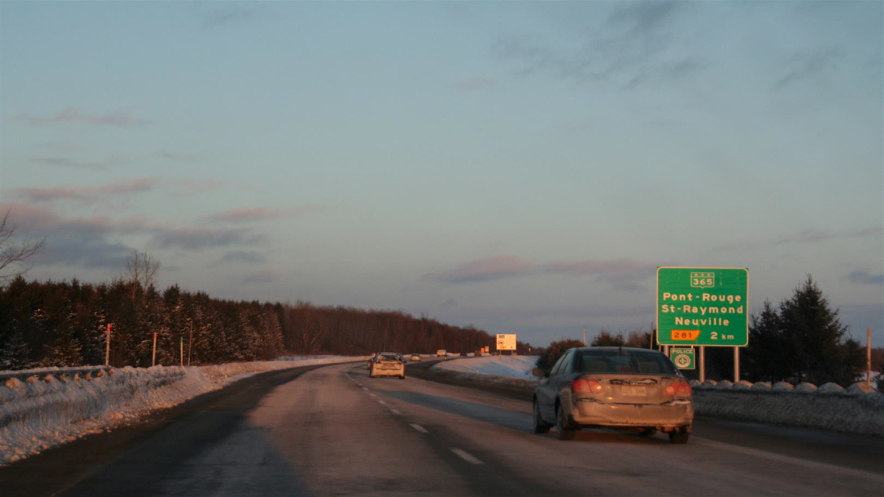 Eine Straße im winterlichen in Quebec mit Verkehrsschildern, die auf die nächsten Orte hinweisen, während die Sonne am Horizont aufgeht