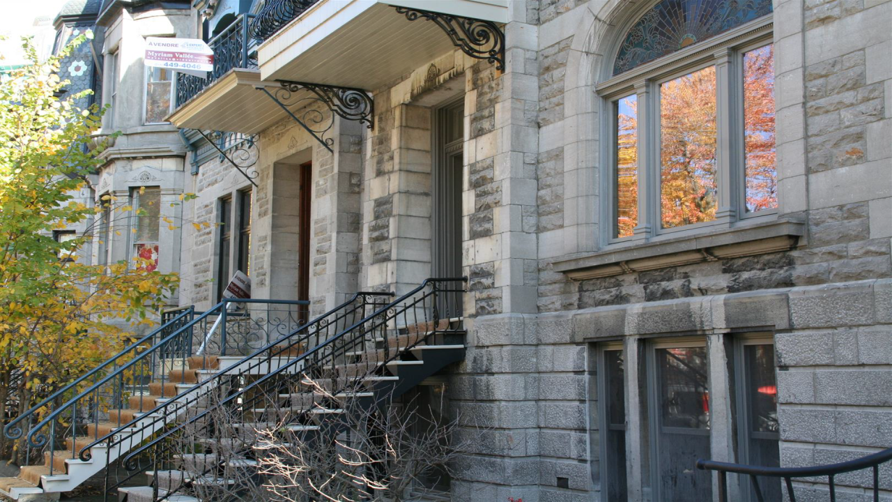 Eine historische Steinfassade mit eleganten Fenstern und einem geschwungenen Geländer, umgeben von herbstlichen Bäumen.
